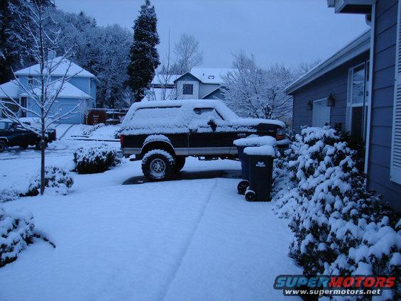 Dark Ford Bronco with a camper shell covered in snow, parked in a driveway next to a house and trash cans