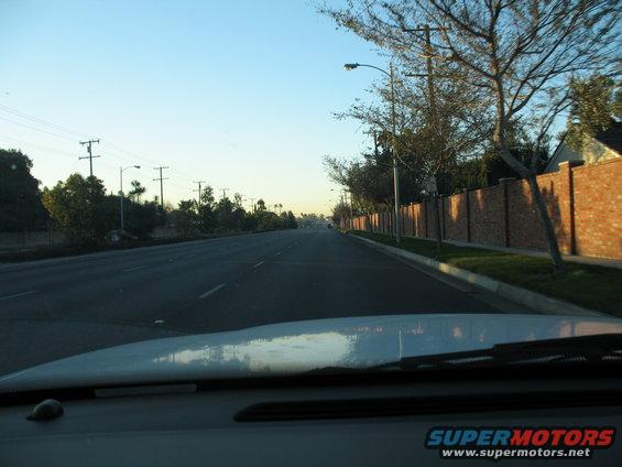 Dashboard and windshield view of a road with a brick wall and trees on the right