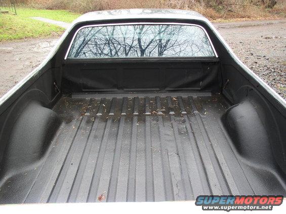 Pickup truck bed with black liner, showing ribbed floor and wheel wells. Trees reflected in rear window