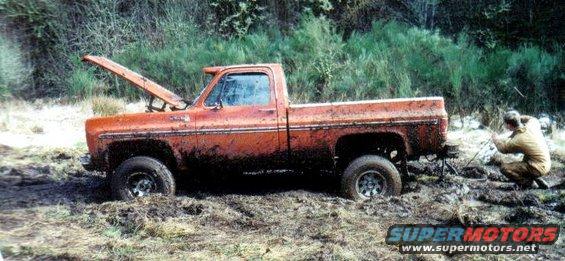 Orange pickup truck stuck in mud with hood open. Person in tan suit is winching it