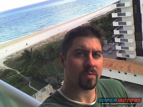 Man takes selfie on balcony with ocean, beach, and hotel in background. "SUPERMOTORS www.supermotors.net" logo visible