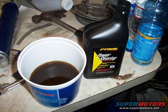Black Pysoil Power Steering fluid bottle next to a white bucket of dark fluid and a hammer