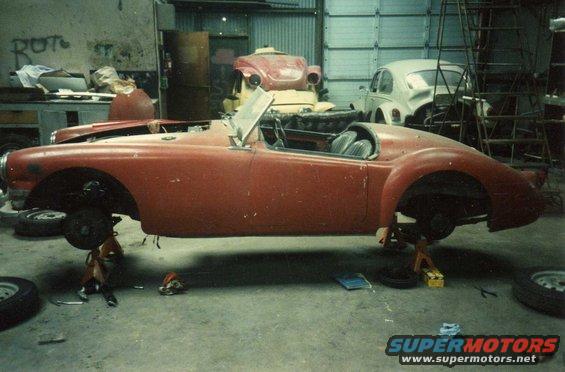 Red 1959 MG A convertible sports car on jack stands in a garage, with other vehicles in the background