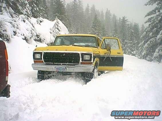 Yellow and black pickup truck with the driver's door open, stuck in deep snow on a wooded road