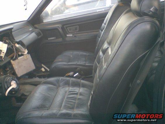 Car interior showing black leather seats and a disassembled dashboard with exposed wires and components