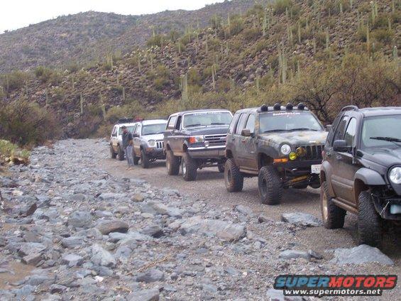 Line of modified SUVs driving on a rocky desert trail with saguaro cacti on the hillsides. Supermotors.net logo visible