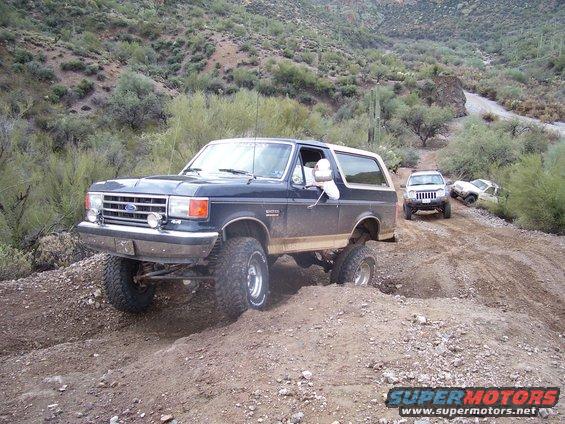 Dark blue Ford Bronco with lifted suspension driving on a dirt trail, other SUVs behind it