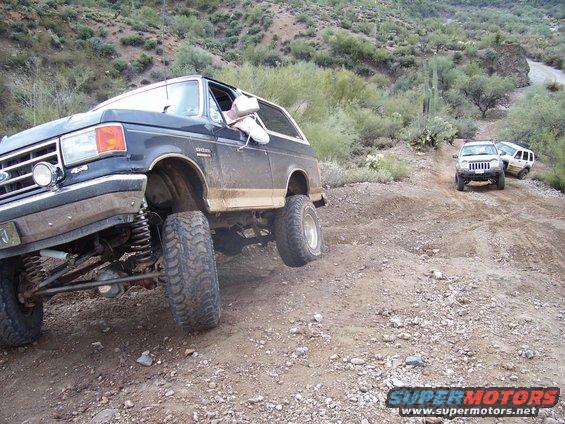 Dark blue Ford Bronco with lifted suspension on a dirt road, driver's window down