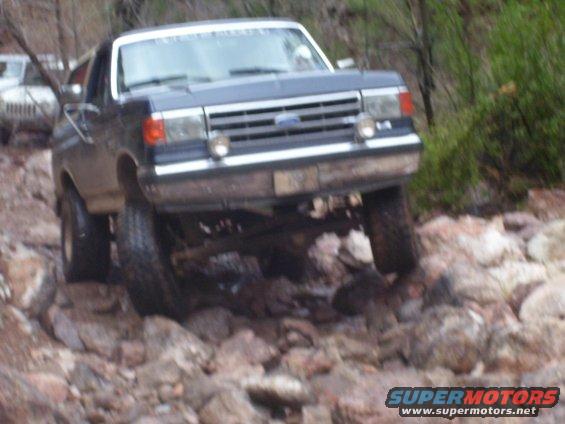 Dark blue Ford Bronco driving over rocks, front 3/4 view. Another white vehicle is visible behind it