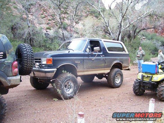 Dark blue Ford Bronco with lifted suspension and large tires parked on dirt road next to another SUV and an ATV