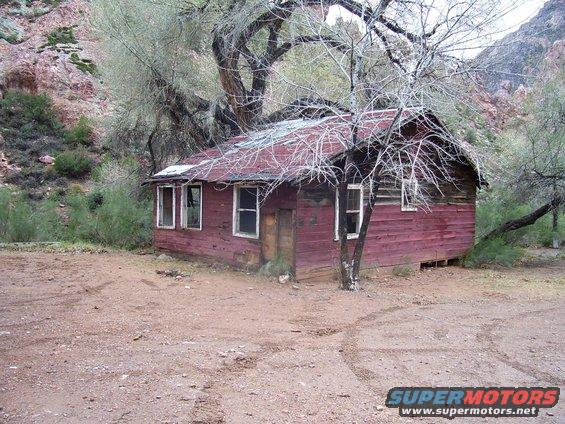 Abandoned red cabin with a damaged roof, surrounded by trees and dry brush. Tire tracks are visible in the dirt