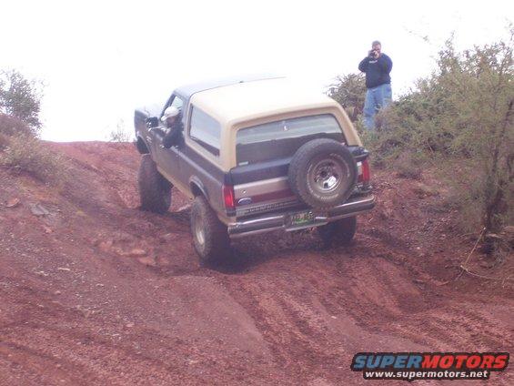 Ford Bronco climbing a dirt hill, spare tire on back. Guy taking photo on hill