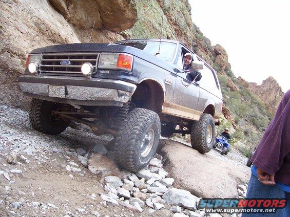 1991 Ford Bronco climbing over rocks on a trail, driver visible. ATV in background