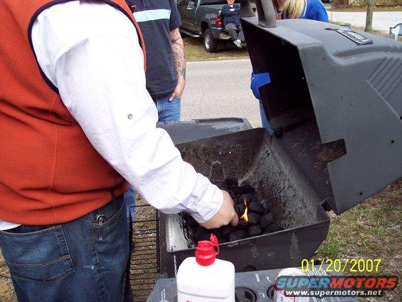 Grill with burning charcoal, people standing around, and a pickup truck in the background