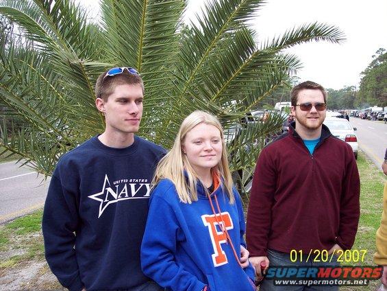 Three people stand outside near palm trees, with cars parked along a road in the background