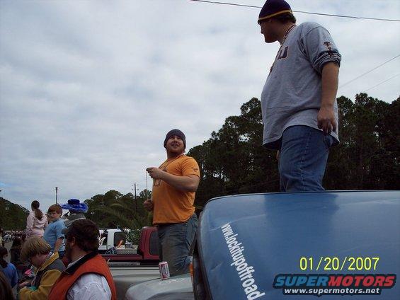Blue vehicle with text 'www.lockitupoffroad' and 'SUPERMOTORS' logo, people standing on it