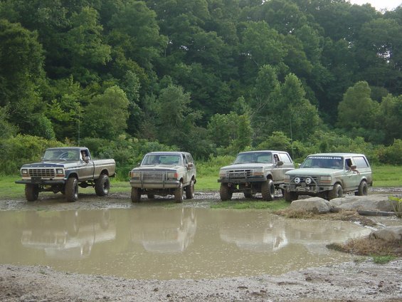 Four lifted Ford trucks and SUVs parked in a muddy field with trees in the background. The first vehicle is a 1979 Ford F-150 pickup
