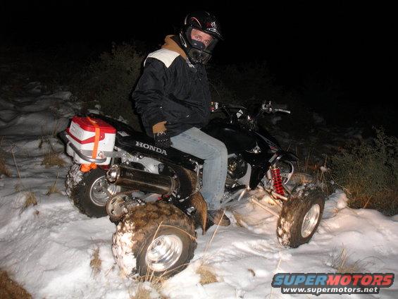 Guy on a black Honda TRX 450 R ATV with a red and white storage box on the back, sitting in the snow at night