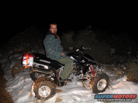 Man sitting on a black 2005 Honda TRX 450 R ATV in the snow at night. The ATV has a red cooler strapped to the back