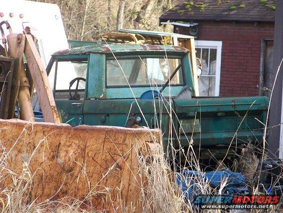 old green pickup truck with rusty roof, parked in weeds next to a brick building