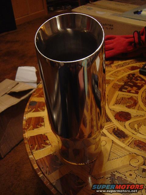 Chrome exhaust tip resting on a table with a wood inlay pattern. Red work gloves and a cardboard box are visible in the background