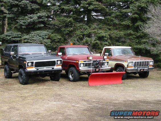 Three trucks parked side-by-side: a black Bronco, a red pickup with a snow plow, and a brown and cream pickup