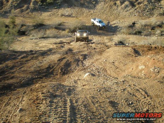 Two trucks driving off-road on a dirt hill with tire tracks visible