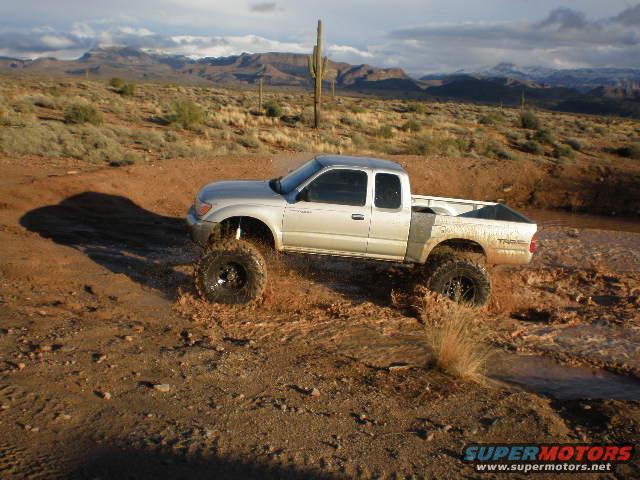 Silver pickup truck with off-road tires splashing through mud in a desert landscape