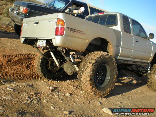 Silver Toyota Tacoma TRD with lifted suspension and large tires, parked behind a black SUV on a dirt trail