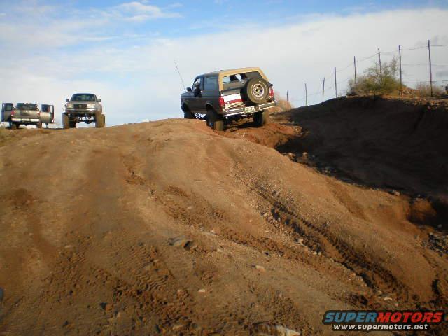 Brown 1987 Ford Bronco driving up a dirt hill, spare tire on rear door, tan top