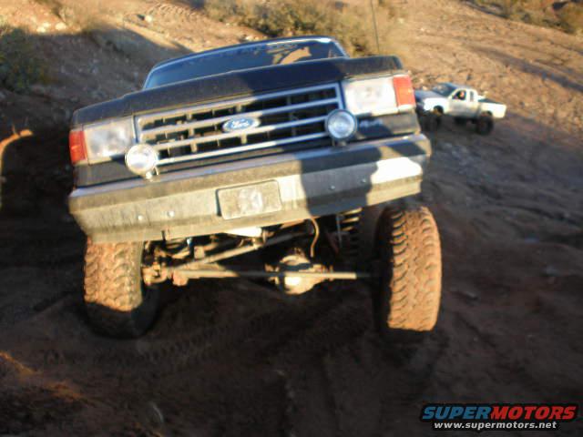 Dark Ford truck front view, lifted suspension, off-road tires, on a dirt hill. Another white truck in background