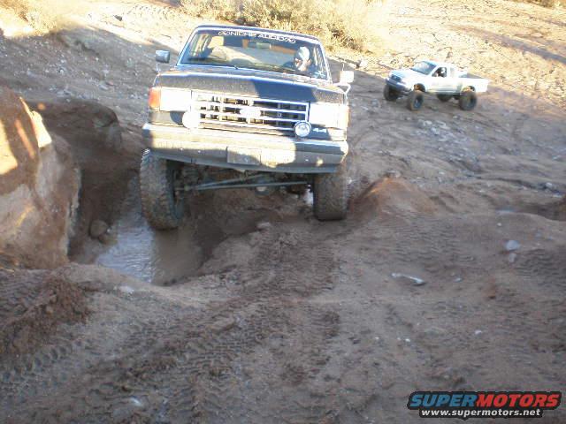 1991 Ford Bronco navigating a muddy trail with a white pickup truck in the background