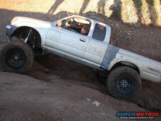 Silver Toyota Tacoma TRD with large tires climbing a dirt hill. Driver visible