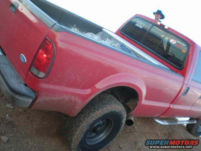 Red pickup truck rear quarter panel, taillight, and bed with a toolbox. Person in cowboy hat visible in cab