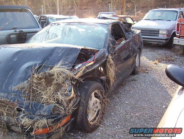 1995 Ford Probe with heavily damaged front end, grass and debris in engine bay, parked in a junkyard with other vehicles