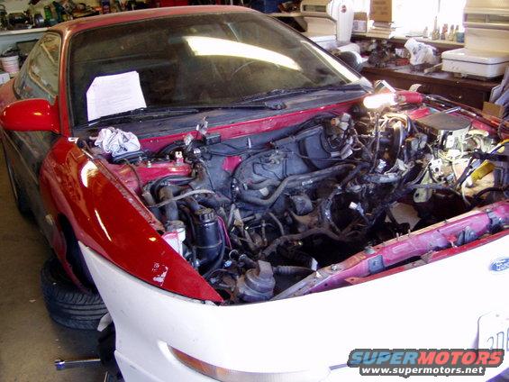red coupe, hood open, engine bay empty, parts on workbench in background