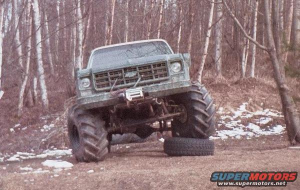 Green monster truck with large tires, winch, and a lifted suspension, parked in a wooded area