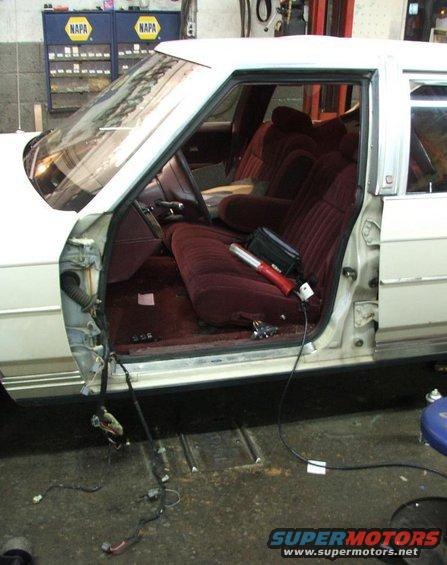 Interior of a white sedan with maroon seats, door removed and wiring exposed