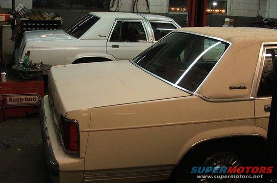 Two sedans, one beige and one white, parked indoors. Beige car in foreground shows rear quarter panel and roofline
