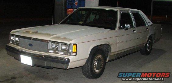 White 1980s Ford sedan, 3/4 front view, parked at a gas station