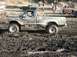 1978 Ford Bronco - A lifted pickup truck navigates a challenging muddy terrain, with spectators and other vehicles in the background.