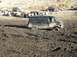 1978 Ford Bronco - A black van is stuck in thick mud, with other vehicles and people visible on higher ground in the background.