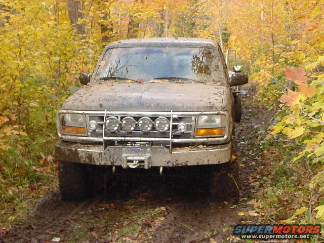 1994 Ford Bronco covered in mud, driving on a wet dirt road with fall foliage