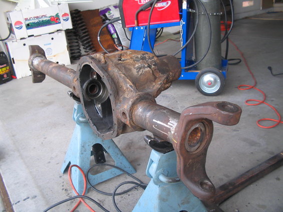 Rusty differential axle housing on jack stands, with welding equipment in background