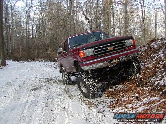 Red 1991 Ford Bronco with lifted suspension and aggressive tires driving on a snowy, wooded trail