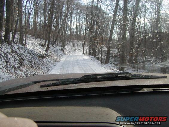 Dashboard and windshield wipers on a vehicle driving on a snowy, tree-lined road