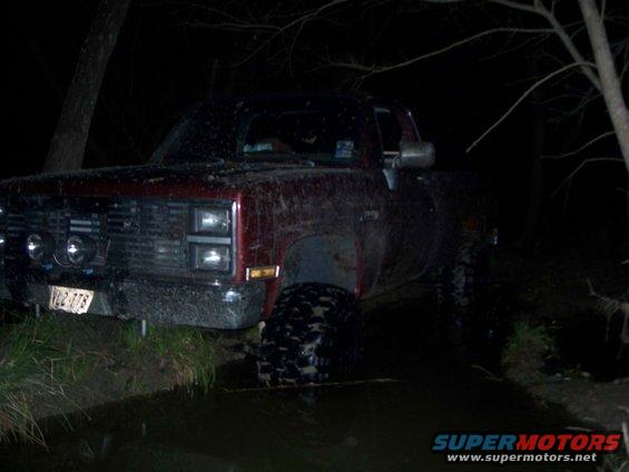 Red pickup truck with large tires partially submerged in water at night. License plate reads VQ2-778
