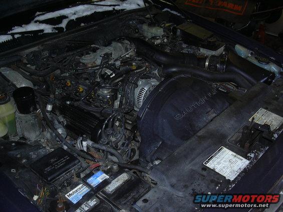 Engine bay with visible battery, air intake, and fan shroud with caution text
