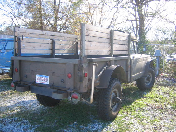 Rear view of olive drab military truck with wooden bed sides. Blue van visible to the left. License plate reads 47H948D
