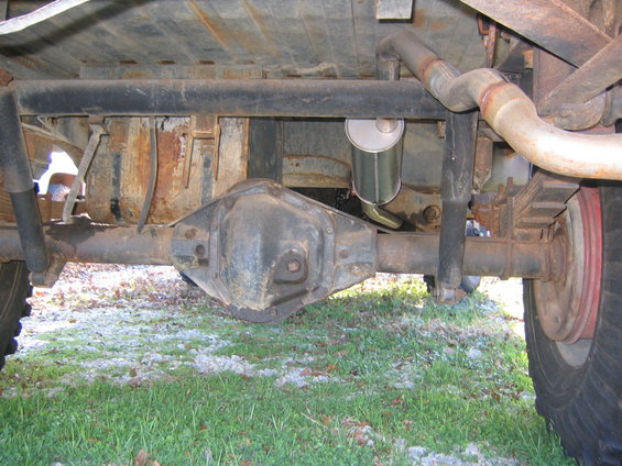 Underside view of a vehicle's rear axle, leaf springs, and exhaust system. Shows rust and wear